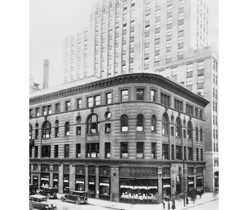 Historic black and white photo of the first Maison Birks store in Montreal, showcasing a prominent corner building with classic architecture. The store features large display windows on the ground floor, filled with goods, beneath a facade of rounded arches and strong stone masonry. Early 20th-century automobiles are parked alongside the curb, and pedestrians can be seen walking by the shopfronts.