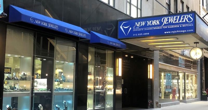 The storefront of New York Jewelers, located on Jewelers Row in Chicago. The exterior features a blue awning with the store's name and logo, and large display windows showcasing various jewelry pieces.